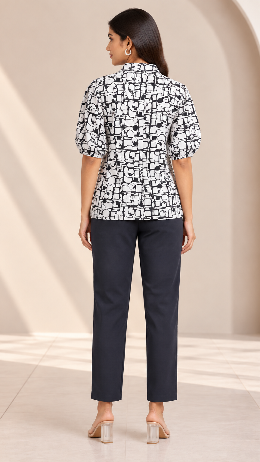 A woman is shown from the back wearing a short-sleeve button-up blouse with a black and white abstract geometric pattern, paired with dark navy trousers and clear high-heeled sandals, set against a minimalist beige architectural background.