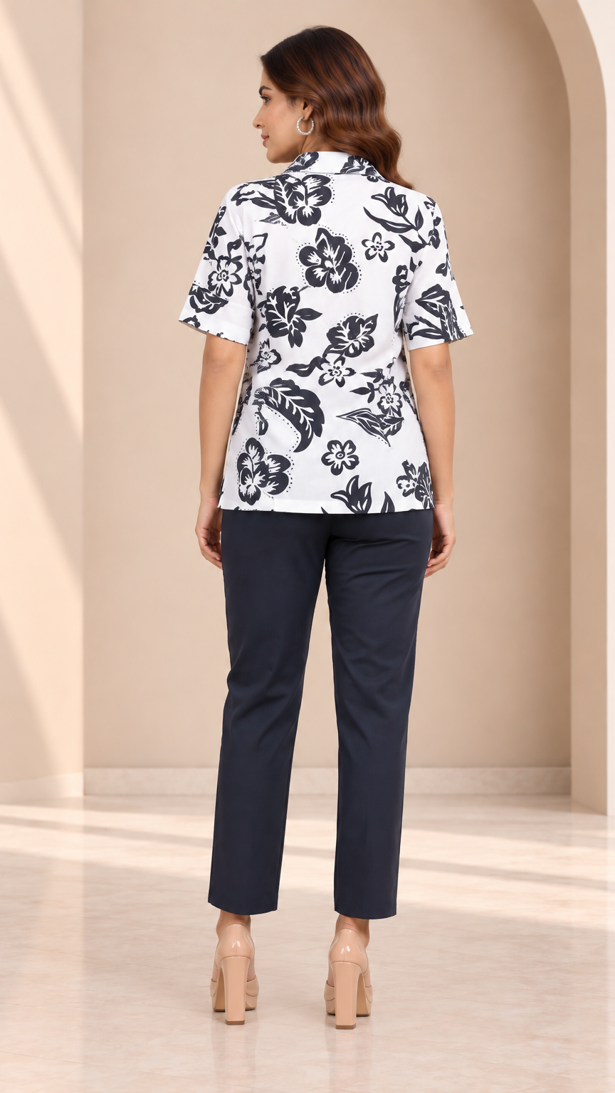 A woman shown from the back wearing a white short-sleeve button-up shirt with a black floral print and dark navy pants, standing in a warm beige interior with soft architectural shadows.