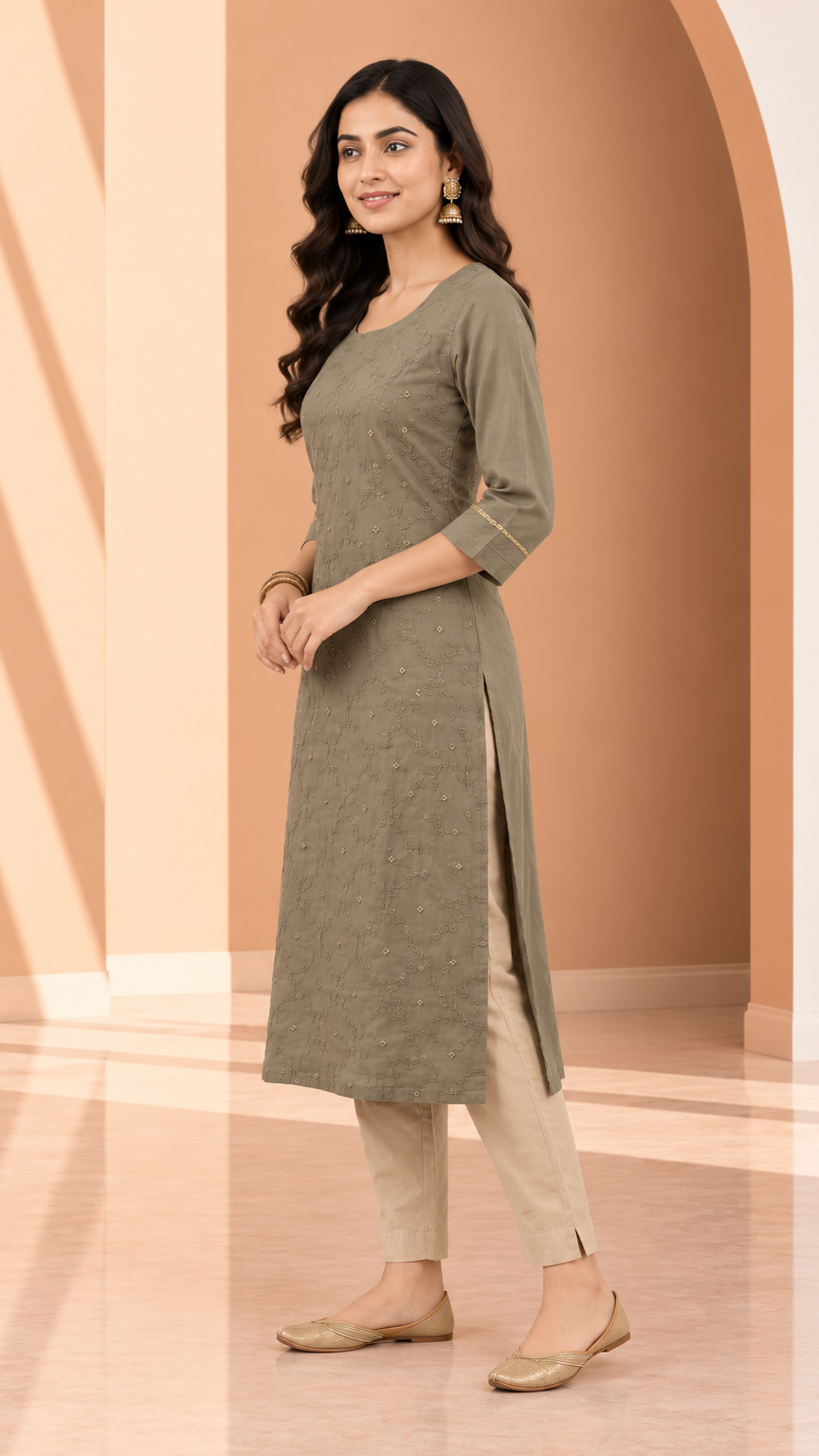 A woman modeling a long olive green kurta with three-quarter sleeves and side slits, paired with beige pants and flat shoes, standing in a warm-toned studio setting with arches.