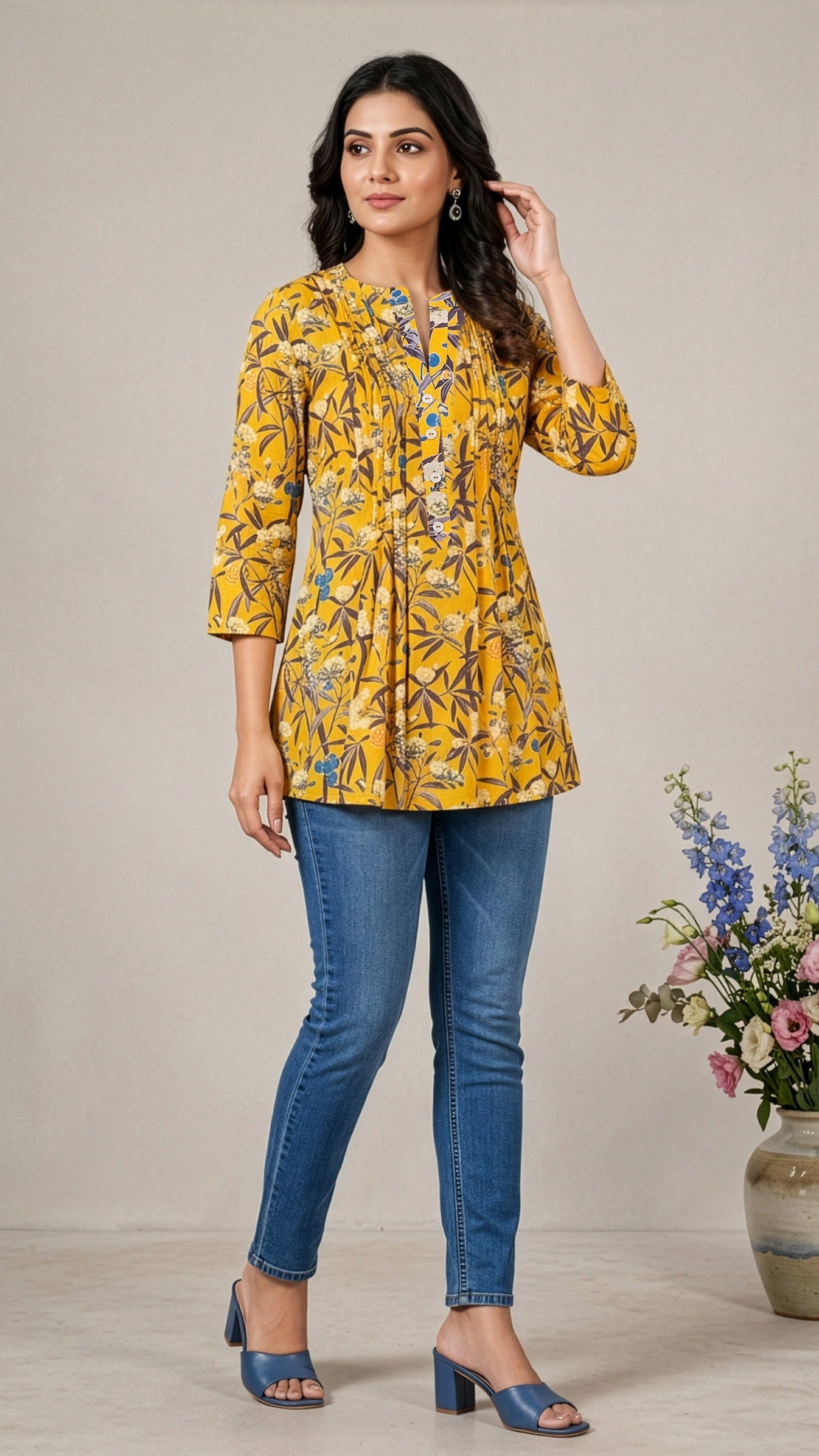 Woman wearing a yellow floral blouse and blue jeans standing next to a vase with flowers.