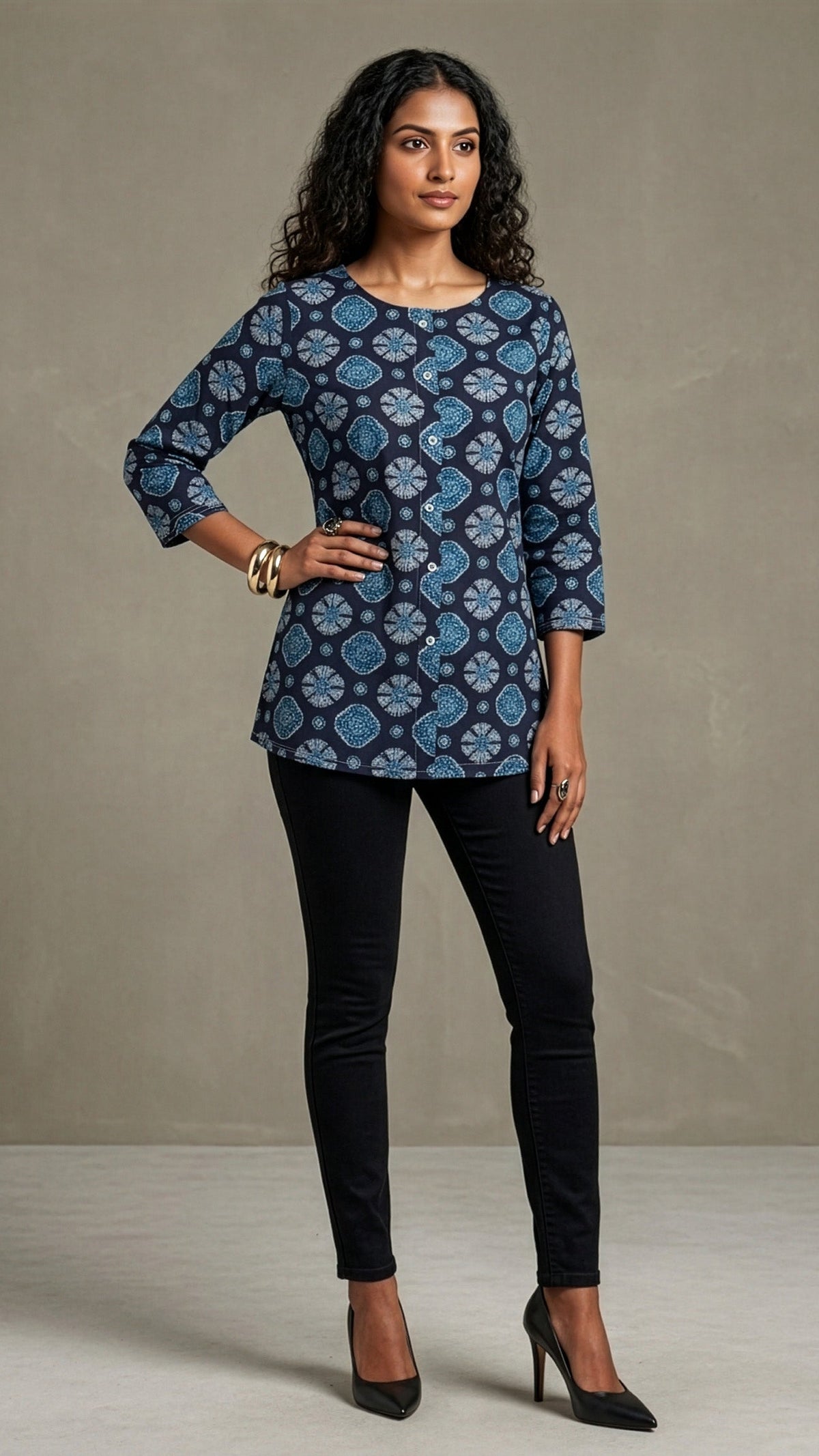 A woman modeling a navy blue three-quarter sleeve top with a blue and light blue paisley pattern, paired with black skinny pants and black heels, standing against a textured gray studio backdrop.