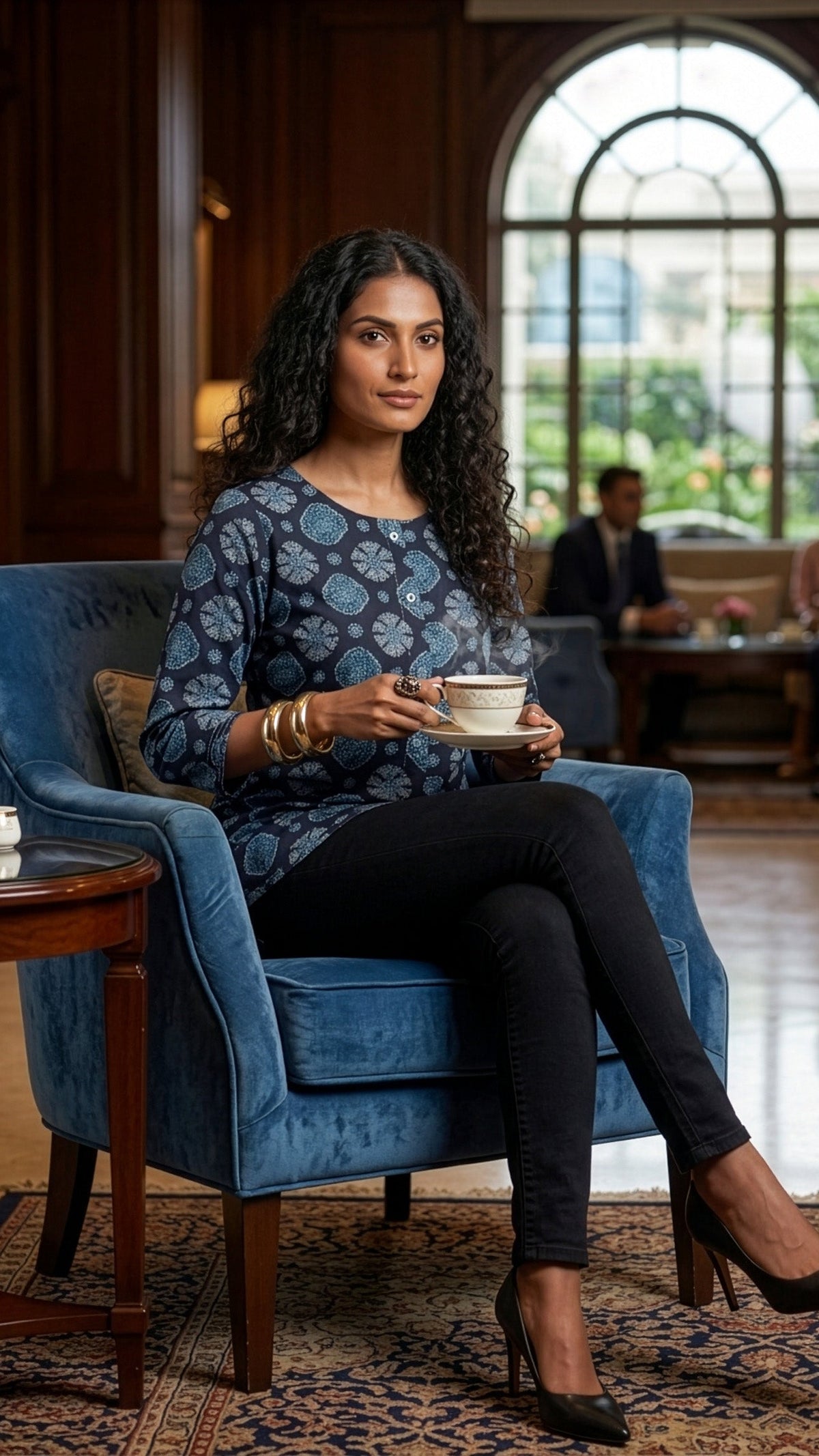 A woman sits in a blue velvet armchair in a polished indoor lounge, holding a white teacup and saucer. She wears a navy patterned long-sleeve top, black pants, and black heels.
