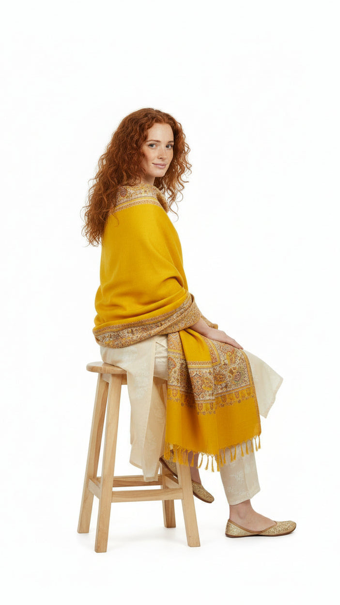 Woman sitting on a stool wearing a yellow traditional outfit with a patterned shawl.