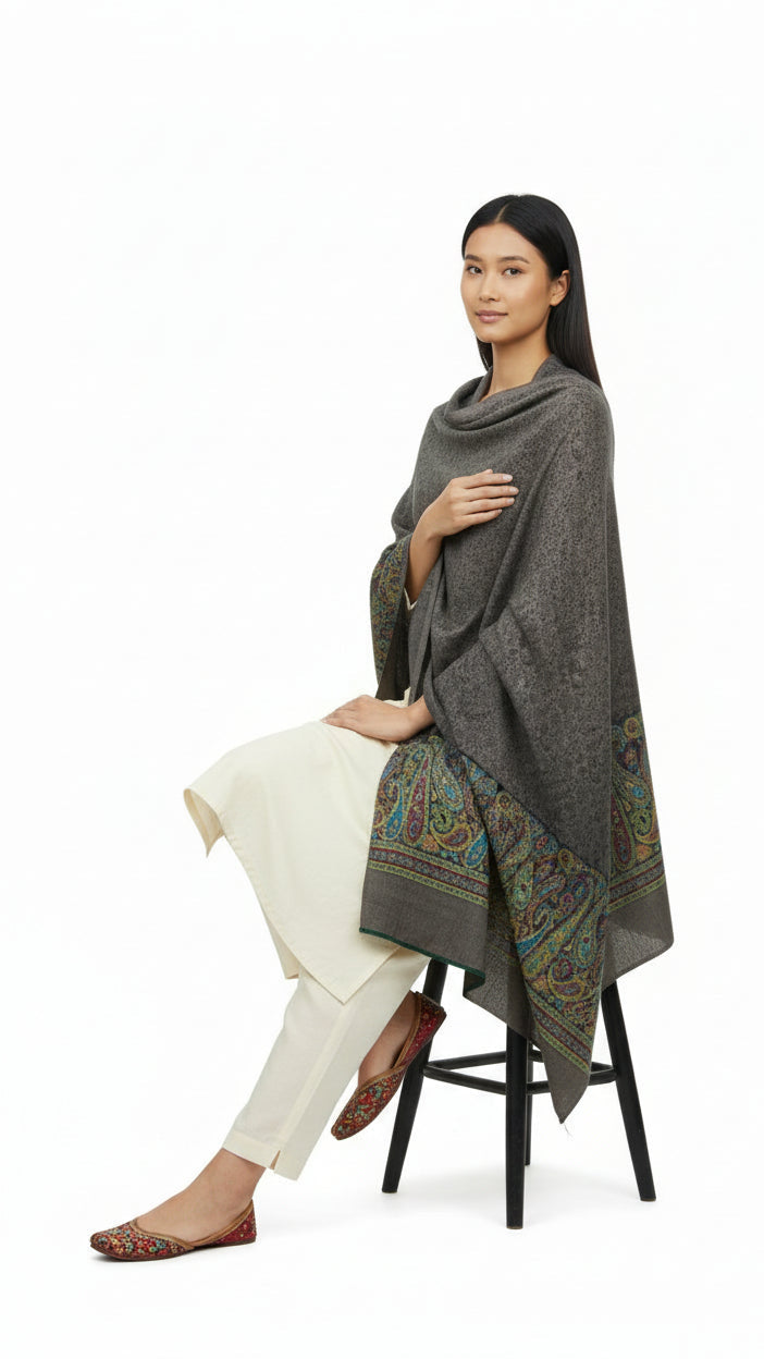 Woman sitting on a stool wearing a gray patterned poncho over a white outfit on a white background
