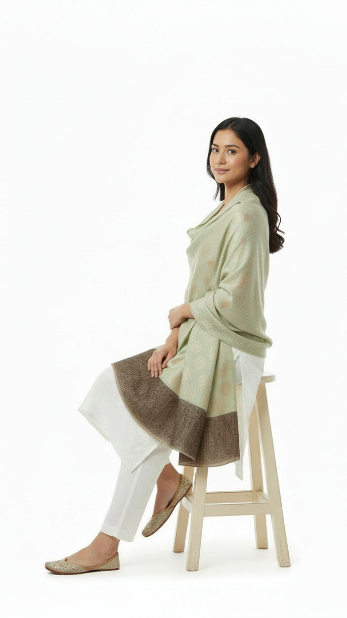 Woman wearing a long green cardigan over a white outfit, sitting on a wooden stool against a white background.