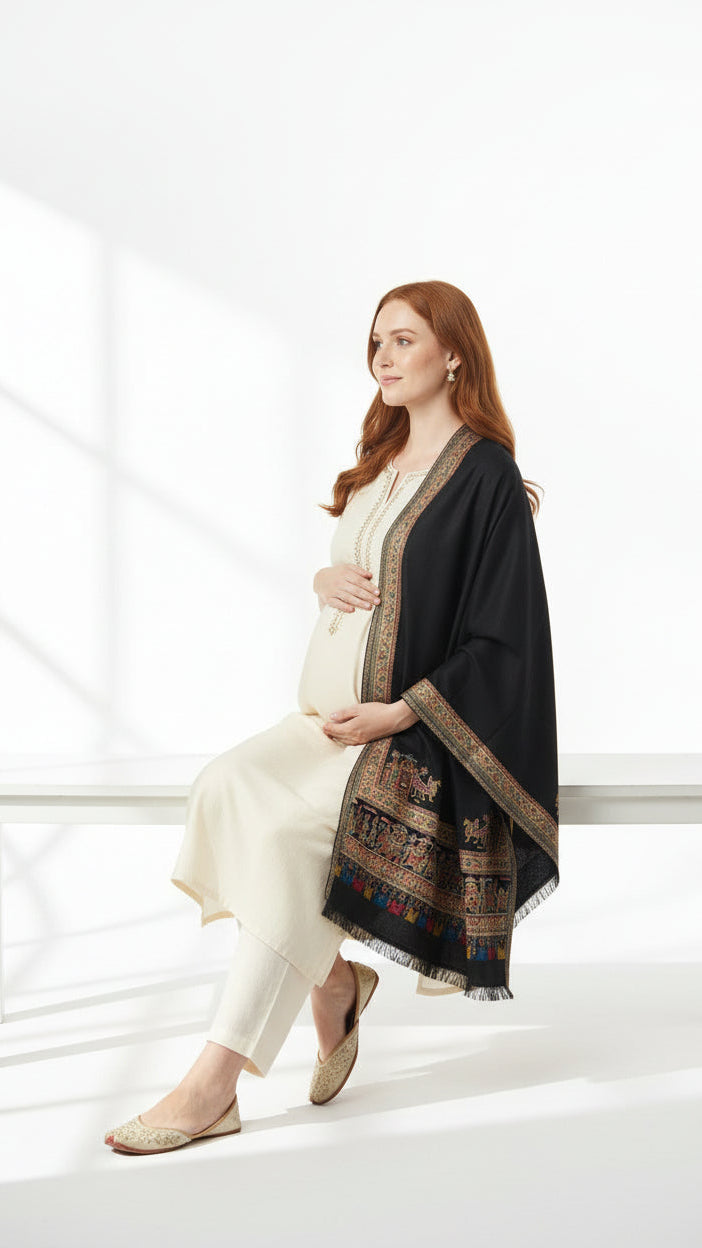 Woman wearing a black shawl with intricate patterns over a white outfit, sitting on a white bench.